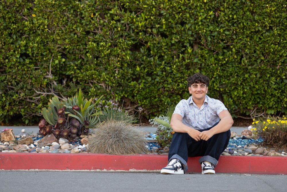 Young man sitting relaxed on a red curb beside desert plants and a green hedge backdrop outdoors.