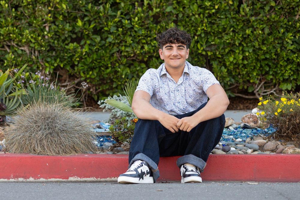 A smiling teenage boy sits on a red curb in front of a garden with succulents, wearing a floral shirt and sneakers.