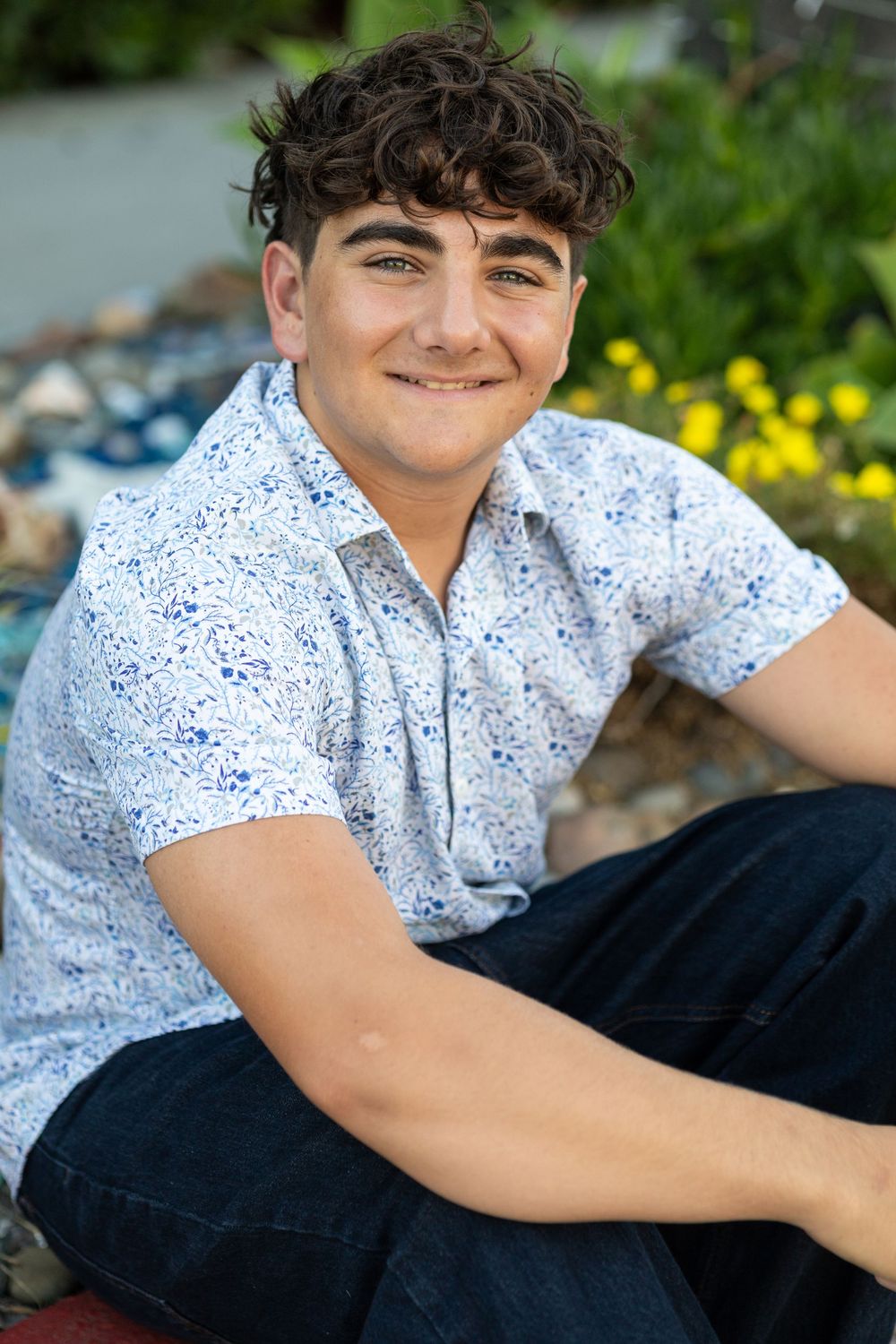 Smiling teenage boy with curly dark hair sitting outdoors in a white blue-speckled shirt and jeans near garden flowers.