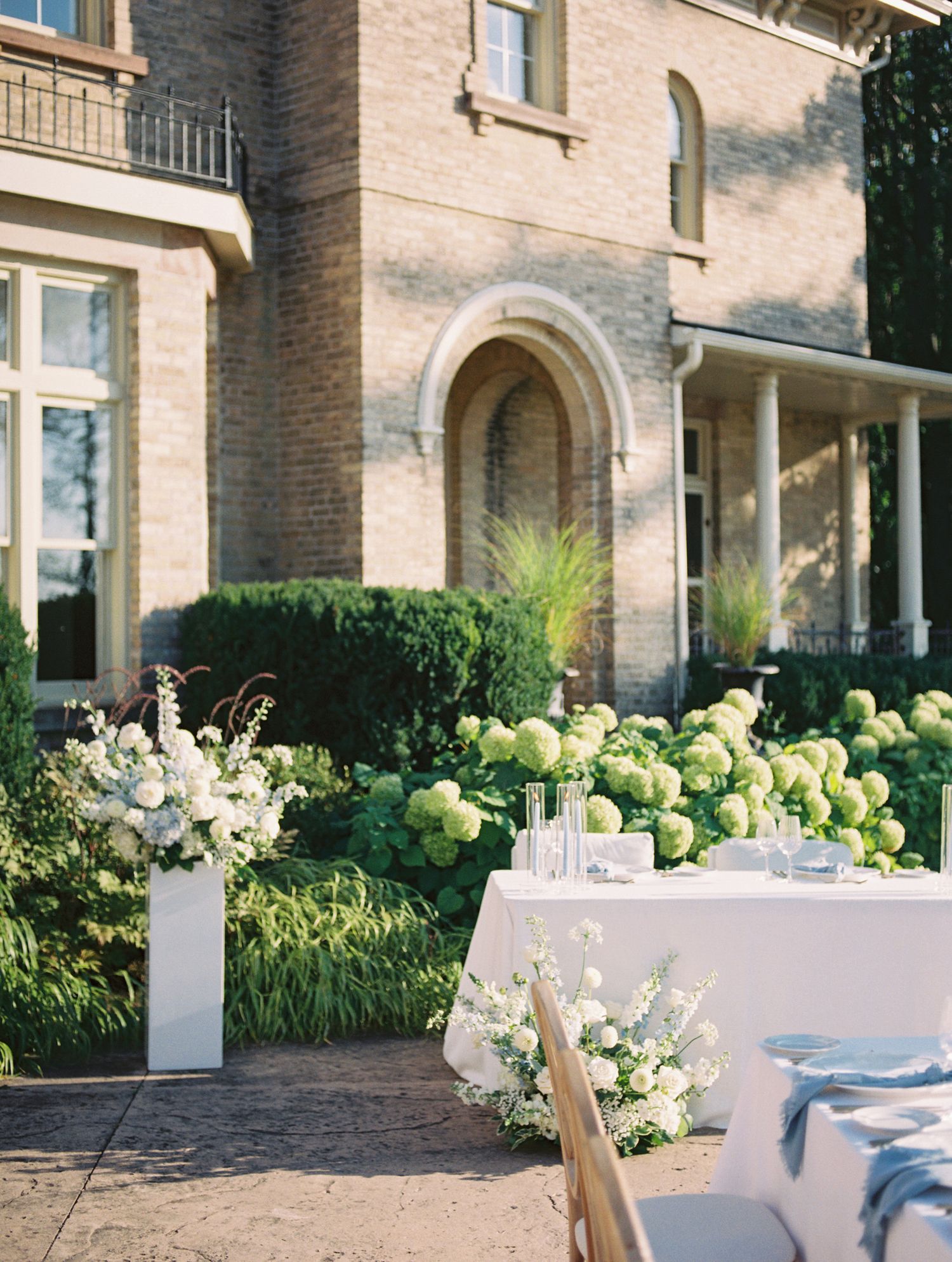 Elegant outdoor wedding reception setup with white floral arrangements and lush green hydrangeas against a classic brick mansion backdrop.