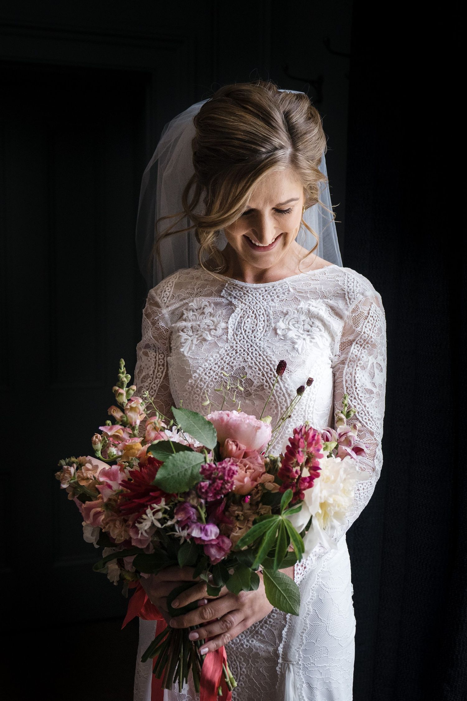 Smiling bride in lace long-sleeve gown and veil holding a pink and red floral bouquet against dark background.