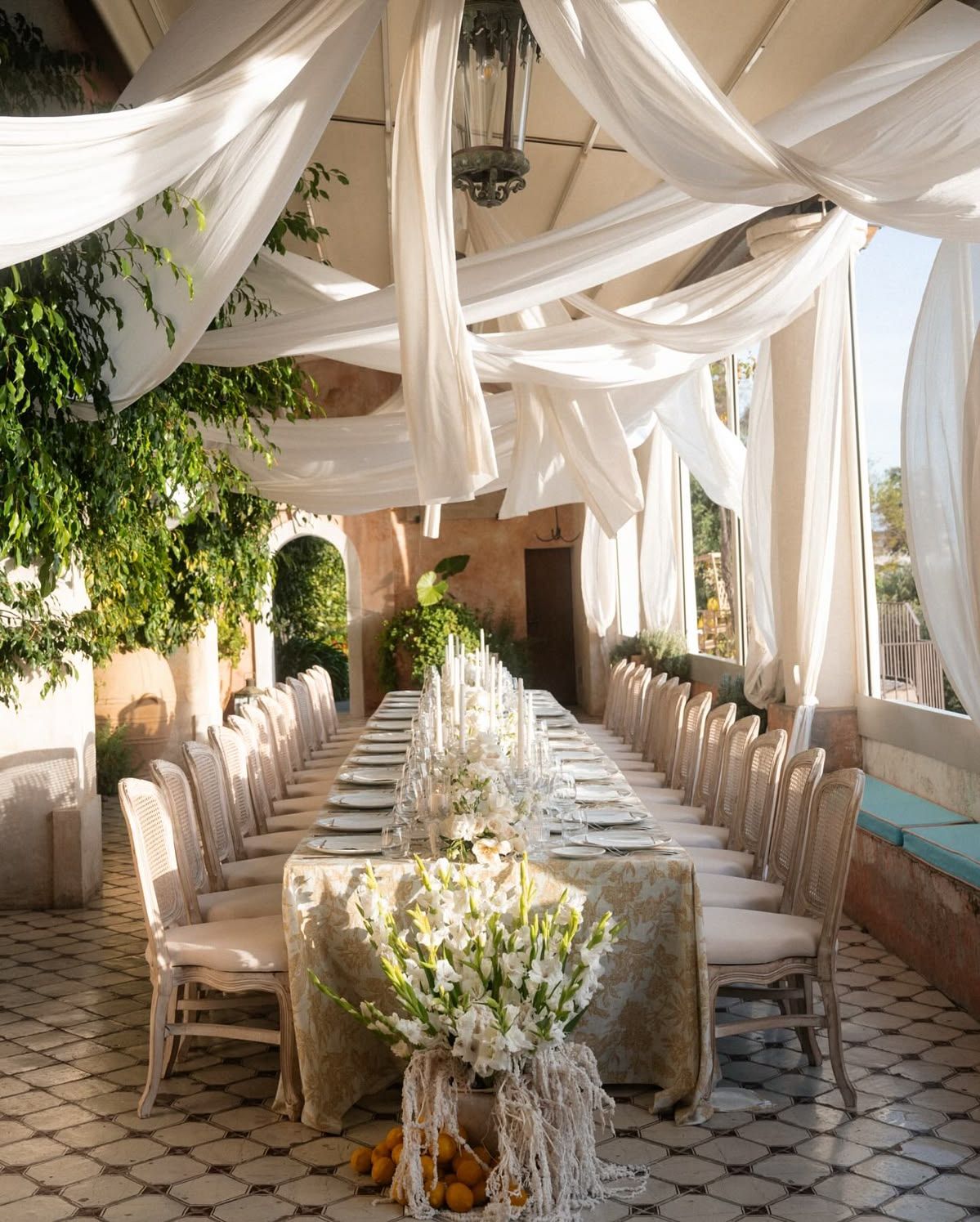 Elegant outdoor wedding reception with white draped fabric ceiling, long dining table, floral centerpieces, and lush greenery.
