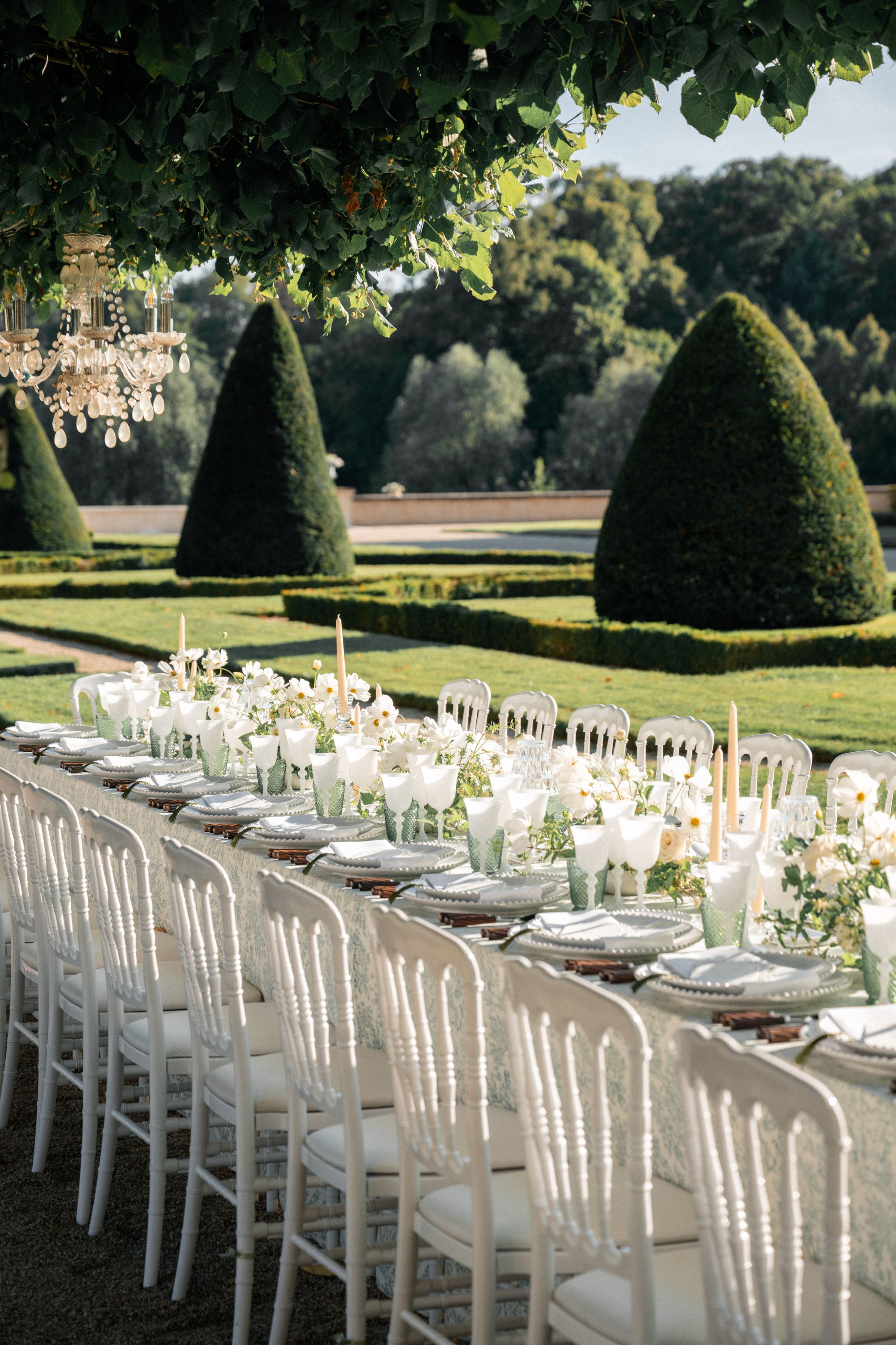 Elegant outdoor wedding reception with white chairs, floral centerpieces, and manicured topiary gardens in the background.