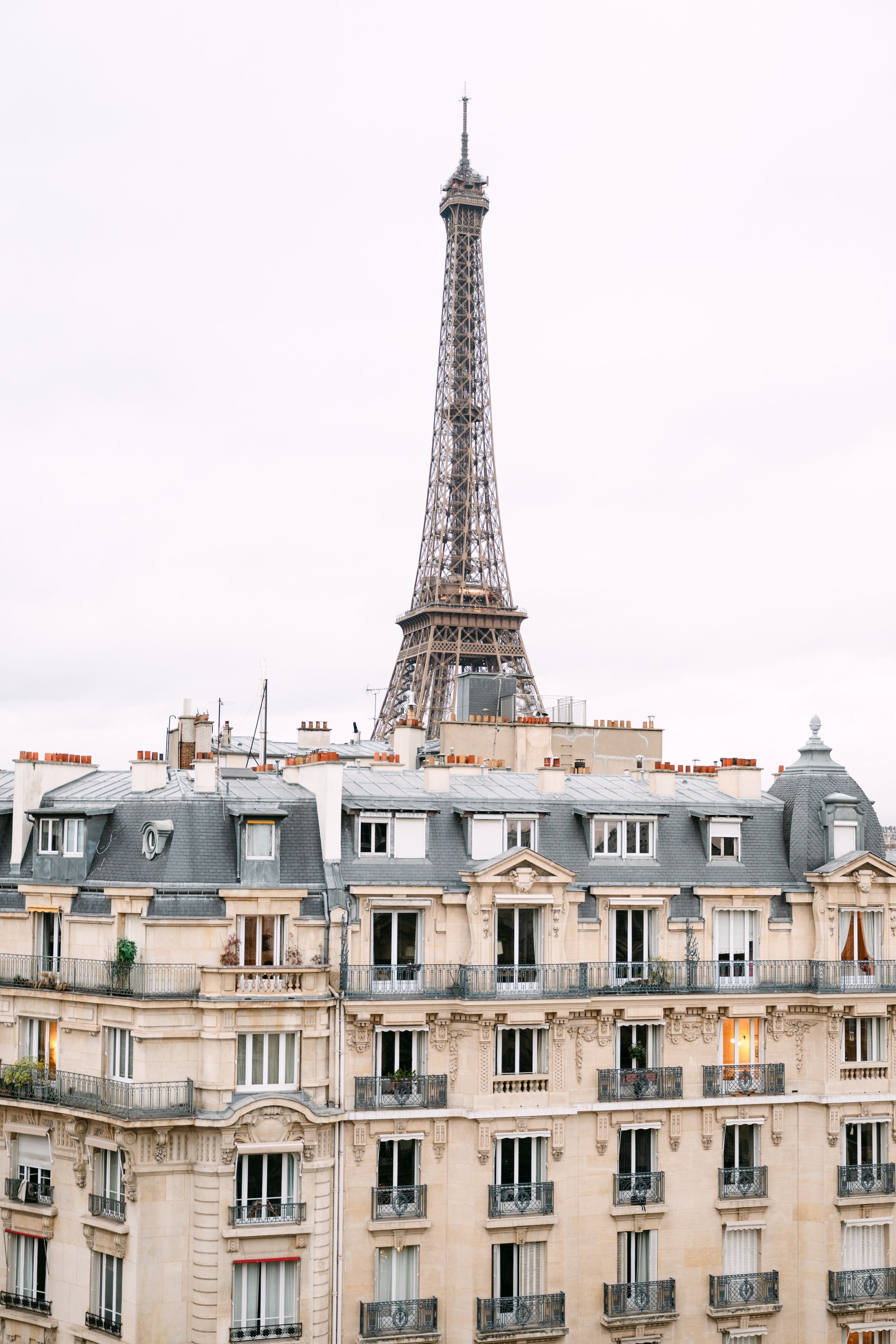 The iconic Eiffel Tower rises above classic Haussmann-style Parisian buildings under an overcast sky.