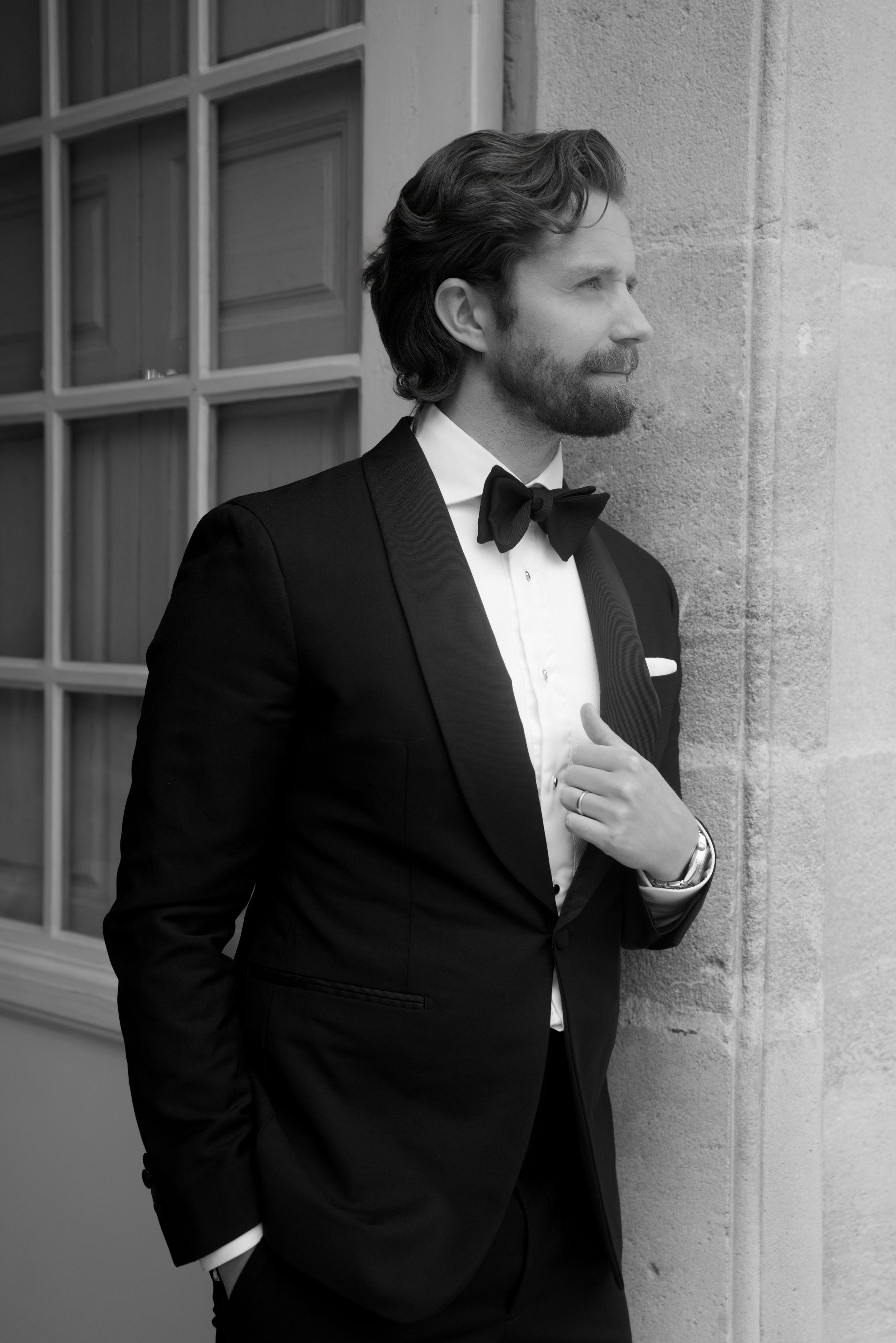 Black and white photo of a bearded man in a tuxedo with bow tie leaning against a stone wall.