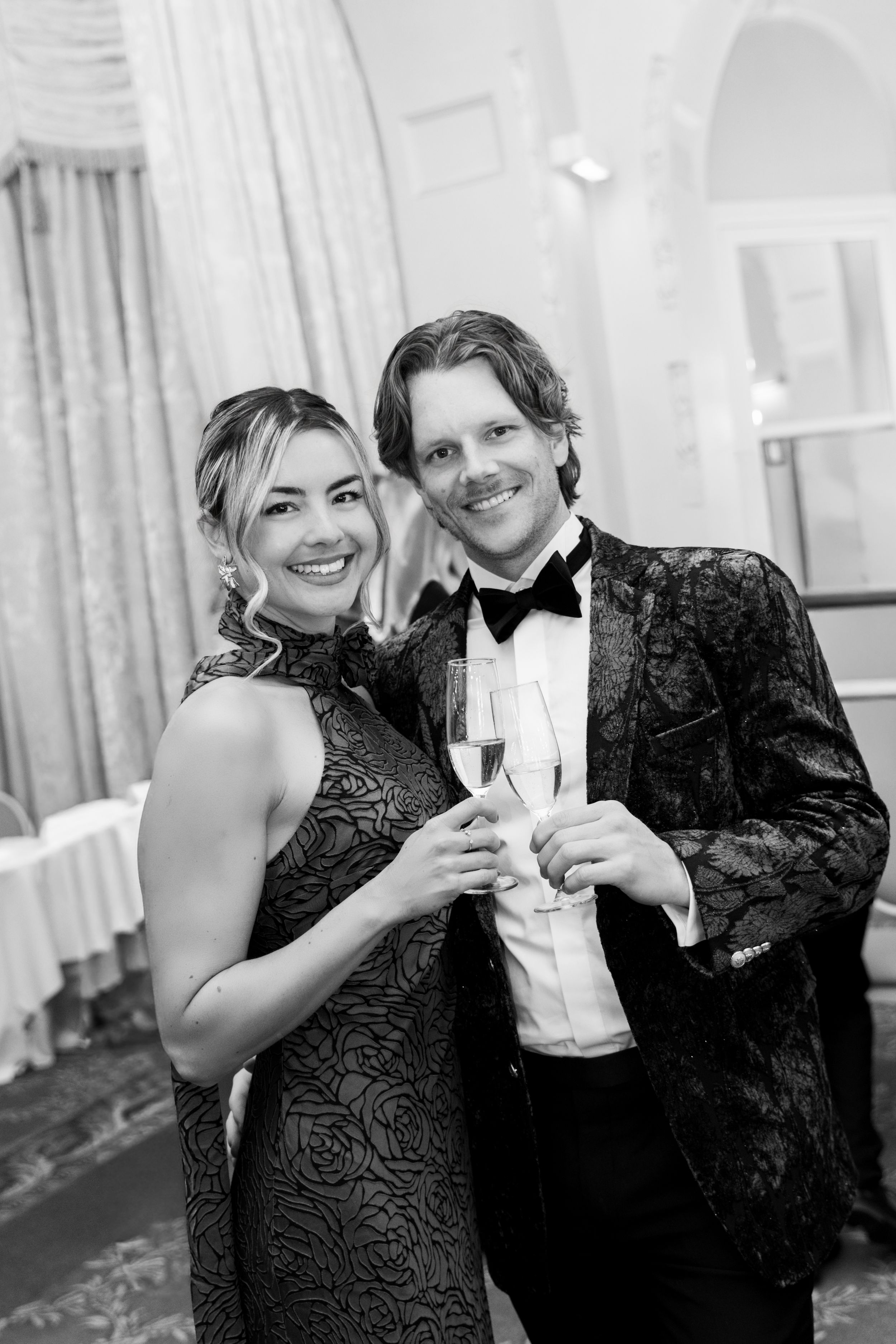 Black and white photo of a smiling couple in formal attire toasting with champagne glasses at an elegant event.