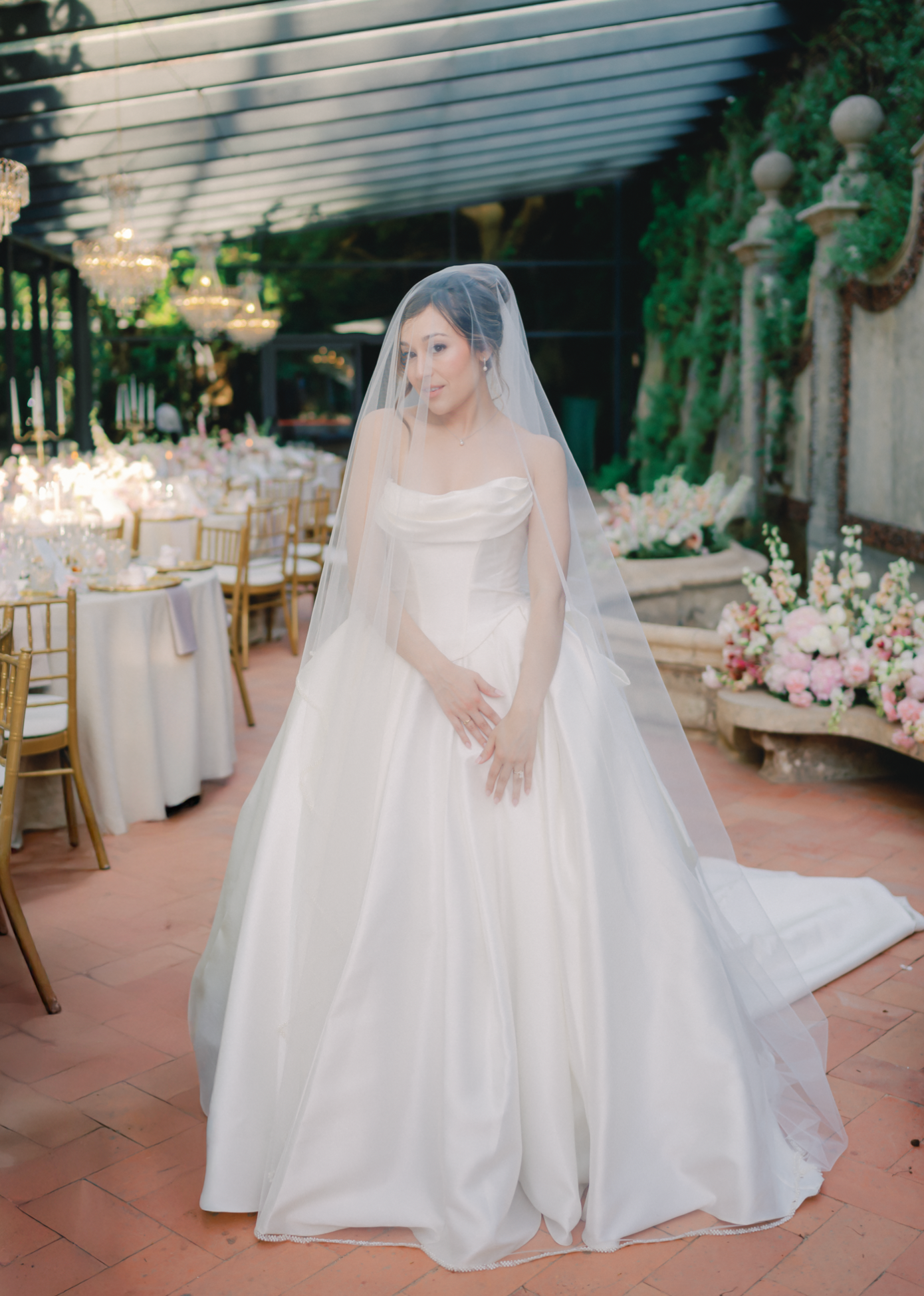 Bride in a strapless ballgown wedding dress with cathedral veil stands in an elegant floral reception venue.