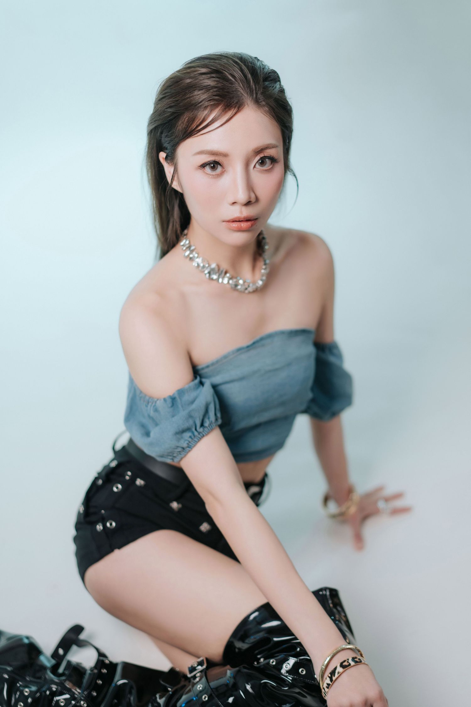 Young Asian woman in off-shoulder denim top, black mini skirt, boots, and pearl necklace posing on white studio floor.