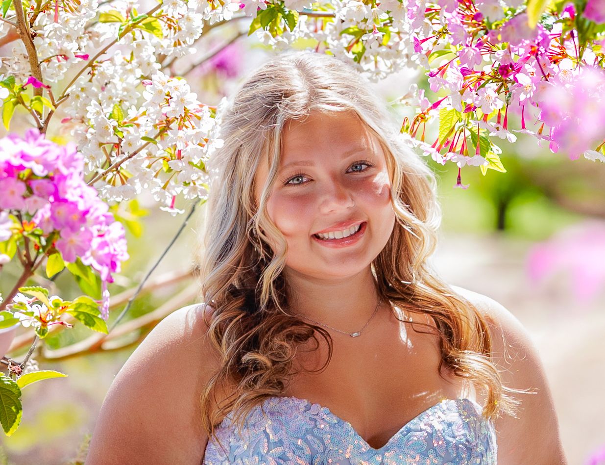 Young blonde woman in blue dress smiling among pink and white flowering trees in a spring garden.