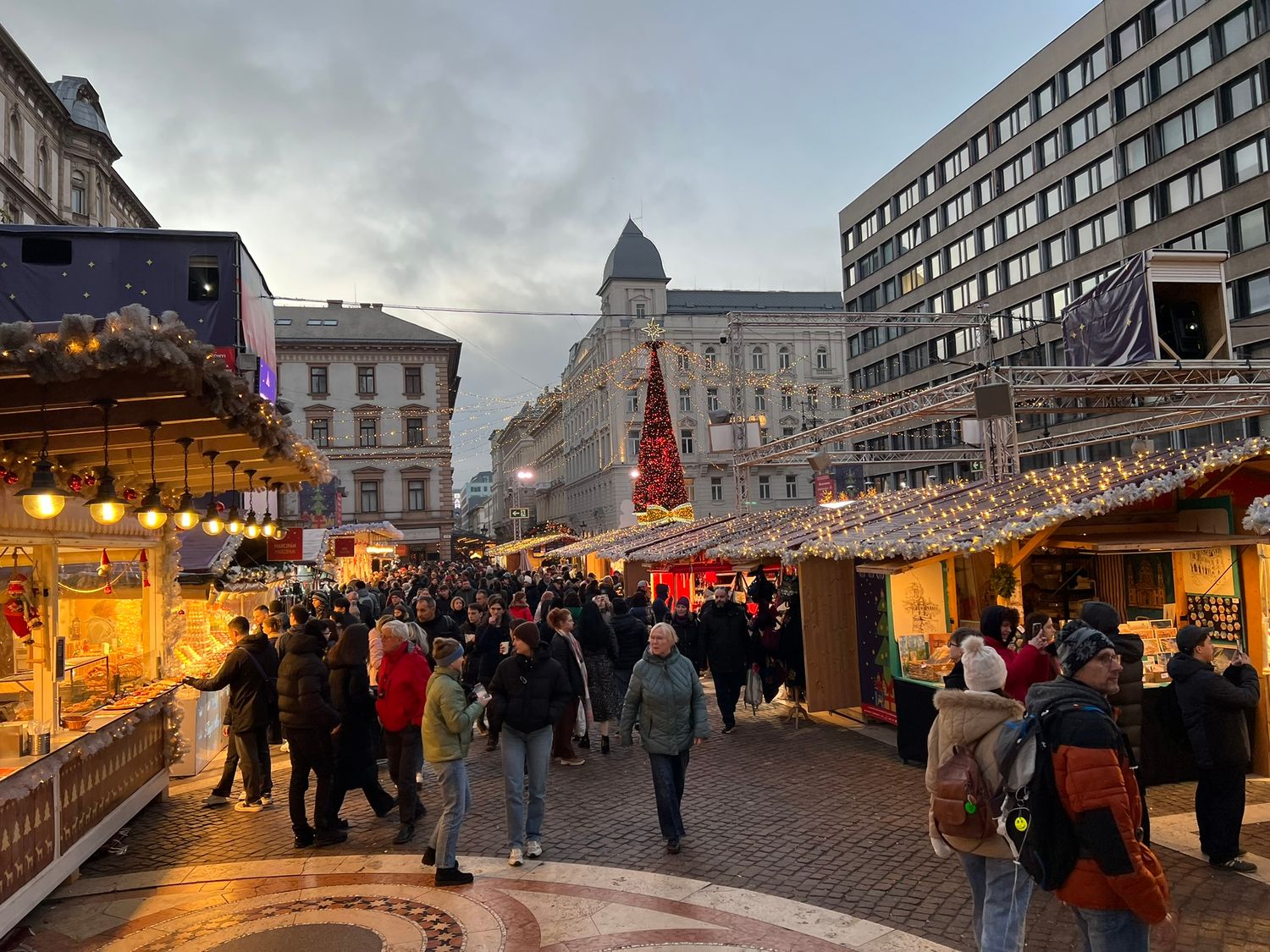 Busy Christmas market with wooden stalls and festive lights filled with holiday shoppers in a European city square at dusk.