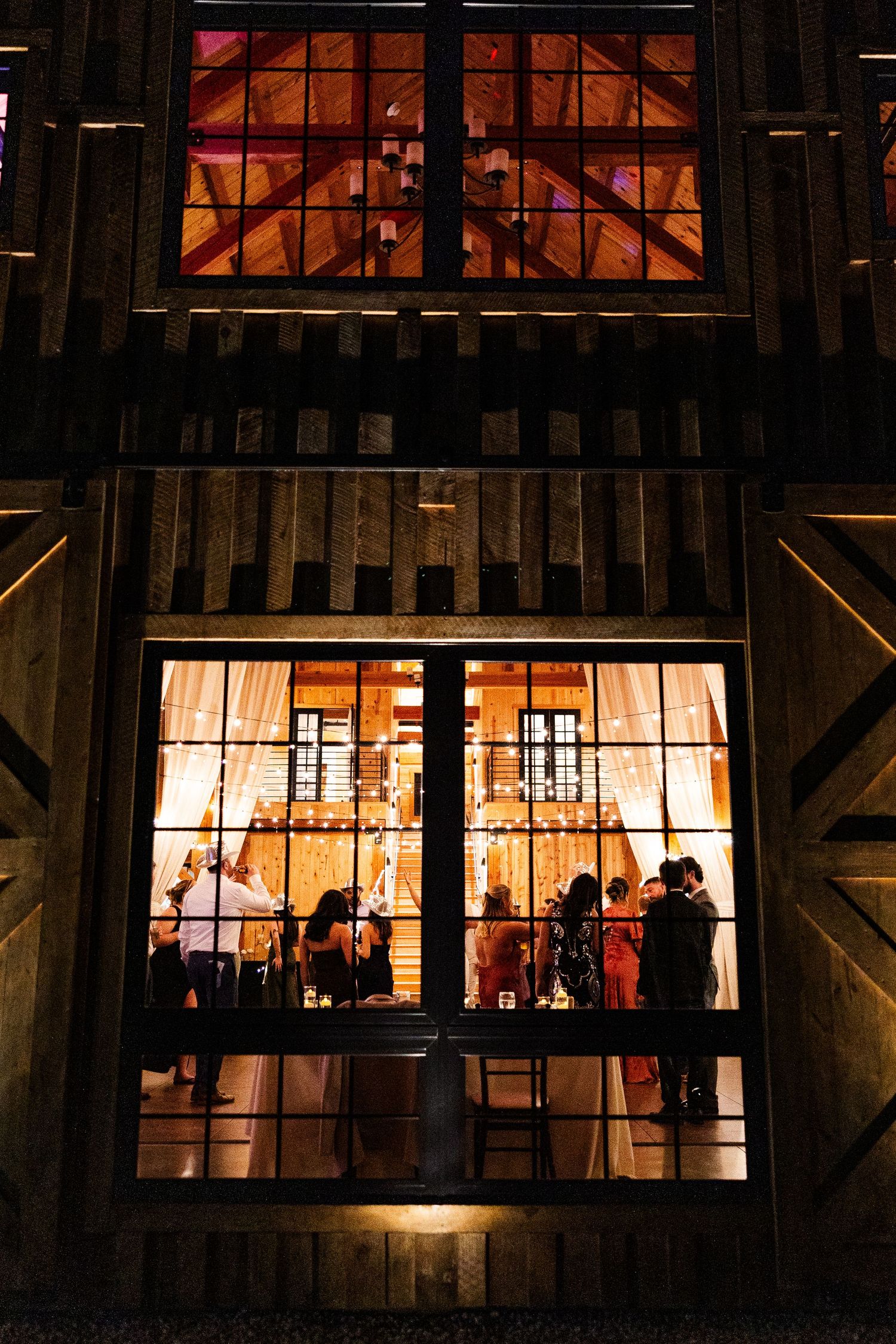 Warm golden light glows through large barn windows at night, silhouetting guests gathered inside a rustic wooden venue.