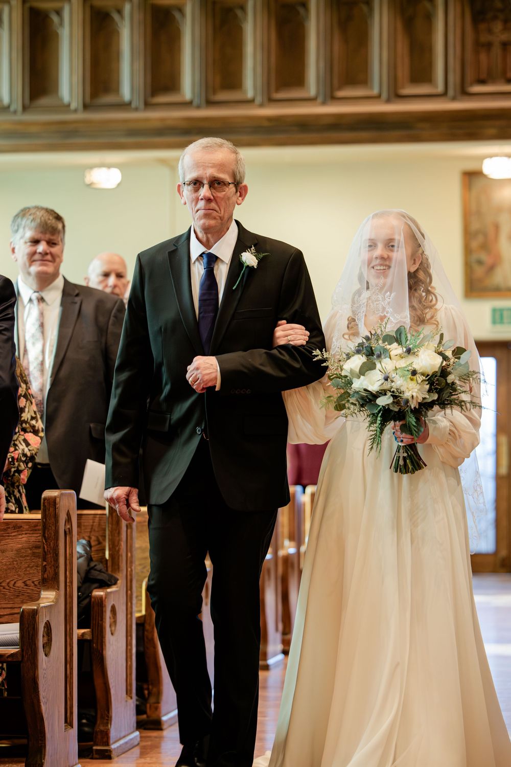 Bride in white gown and veil walks down church aisle with escort holding bouquet of white and green flowers.