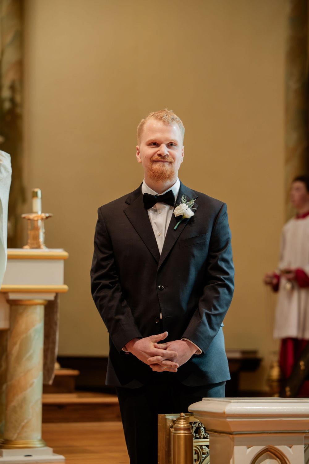 Groom in black tuxedo with white boutonniere smiling at church altar awaiting bride during wedding ceremony.