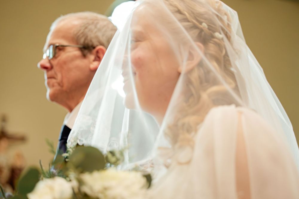 Bride in white veil holding bouquet stands beside older man in suit during wedding ceremony in church.