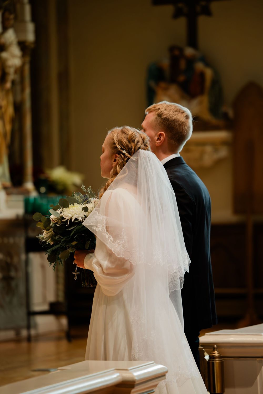 Bride and groom standing at church altar during wedding ceremony, bride holding white floral bouquet with veil.