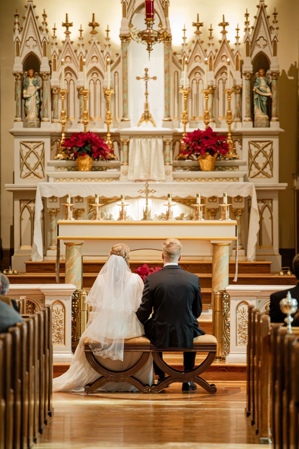 Bride and groom kneeling at ornate white and gold church altar with red poinsettias during wedding ceremony.