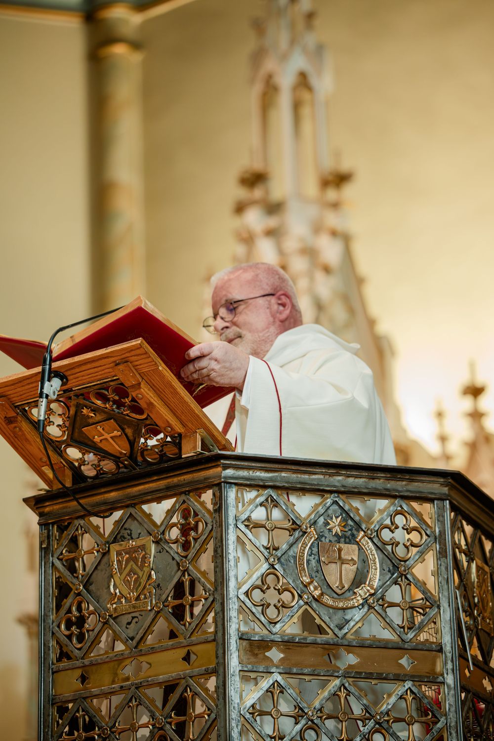Elderly priest in white vestments delivering a sermon at an ornate Gothic church pulpit with red book.