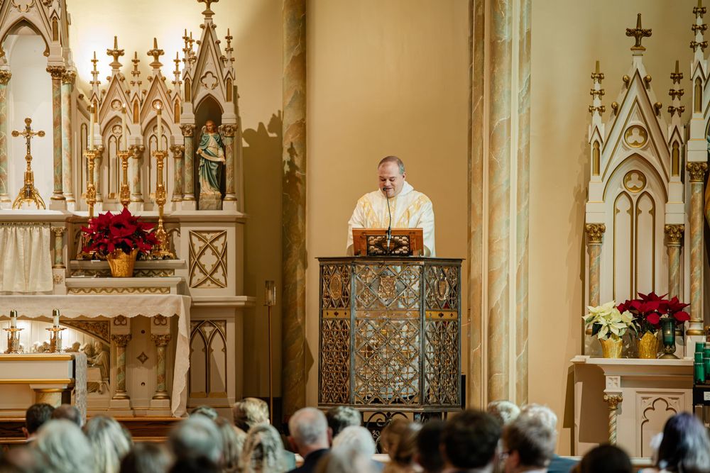 Priest delivering a sermon at an ornate Catholic church altar adorned with gold details and red poinsettias.