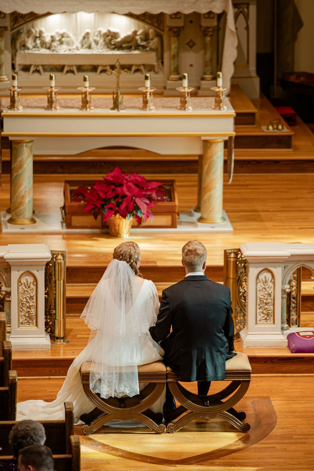 Bride and groom kneeling together at the altar during a Catholic church wedding ceremony with poinsettia floral arrangement.