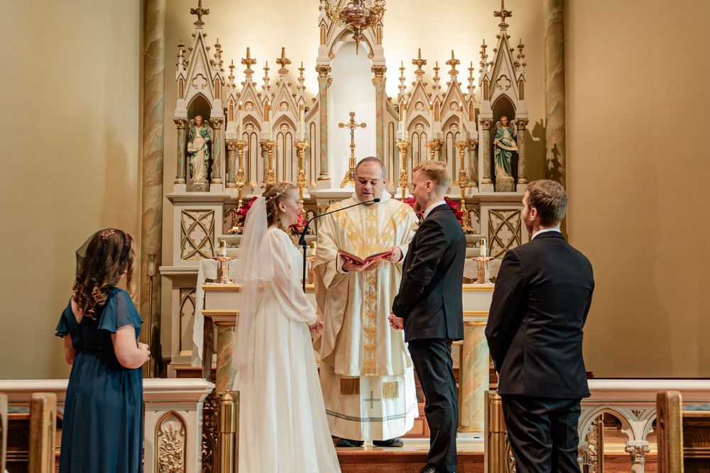Bride and groom exchange vows at ornate church altar with officiant and wedding party witnesses.