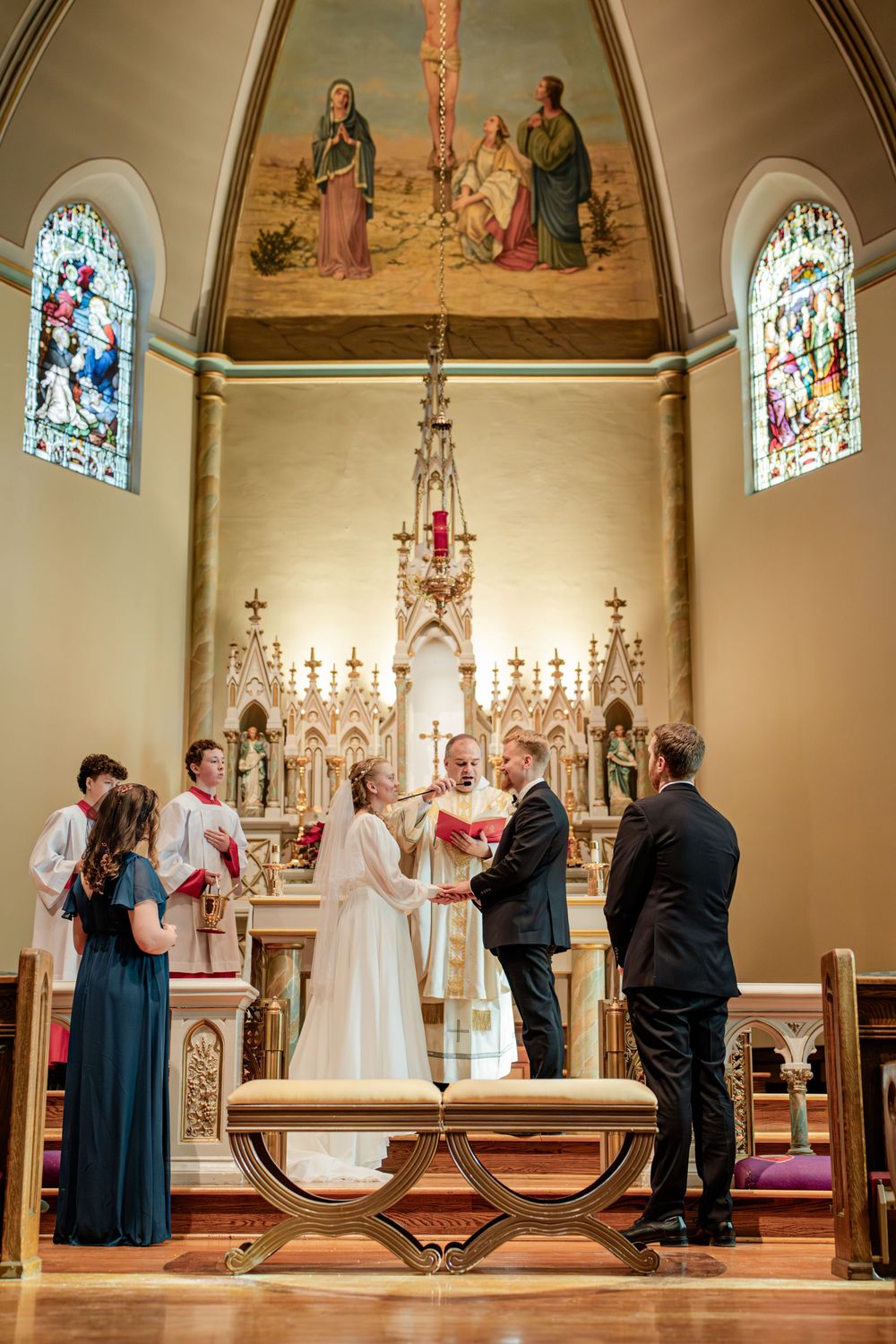 Bride and groom exchange vows at ornate church altar with stained glass windows and religious paintings.