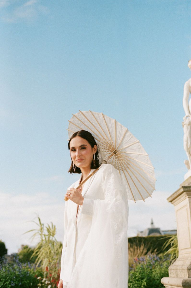 Woman in white outfit holding a cream paper parasol umbrella outdoors near classical sculpture on a sunny day.