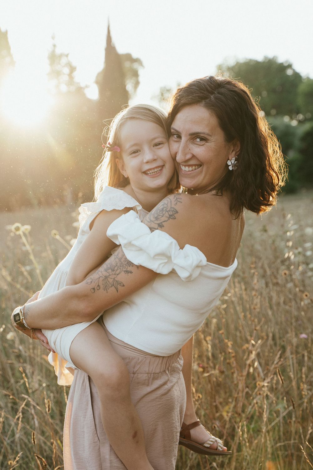 Woman holding smiling girl in sunlit field at golden hour, both wearing white tops outdoors.