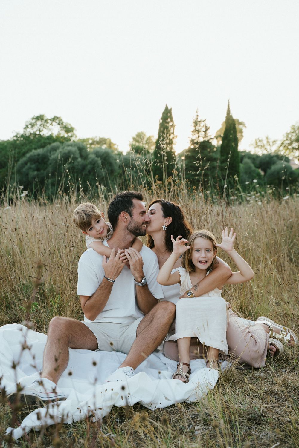 Family of four sitting in golden field, parents kissing while two young children smile and play around them.