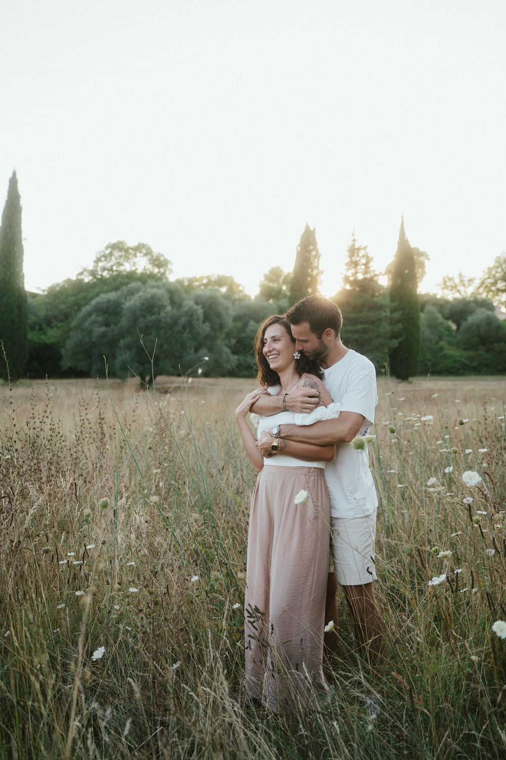 Couple embracing in a sunlit wildflower field at golden hour, woman in pink dress smiling, man in white t-shirt.