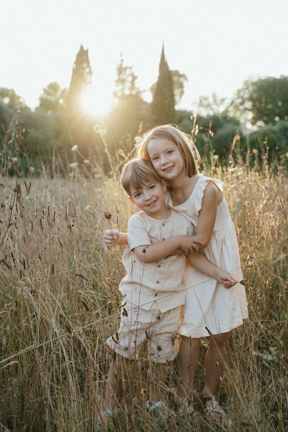 Two children hugging in a golden field at sunset, wearing cream outfits, with warm sunlight glowing through tall grass.