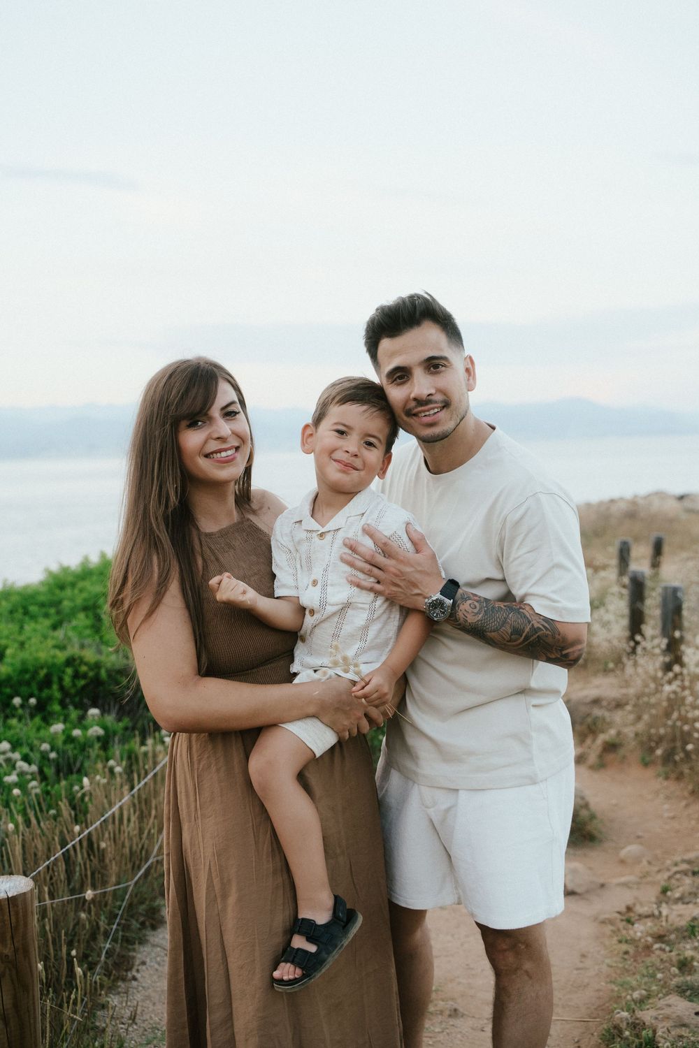 A happy young family with a tattooed father, mother in brown dress, and young son pose outdoors near the ocean.