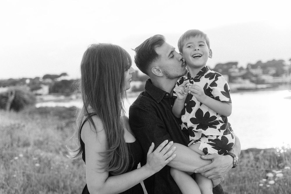 Black and white photo of a couple kissing their smiling toddler outdoors near a scenic waterfront.
