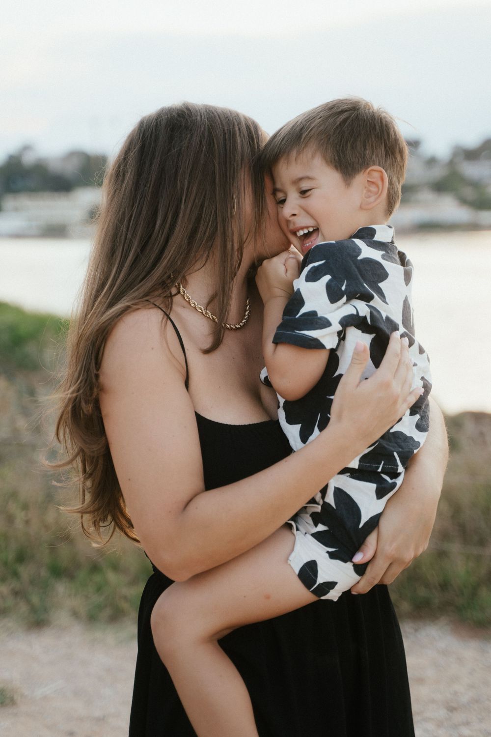 A mother in a black dress holds her laughing toddler boy in a floral outfit near a beach.