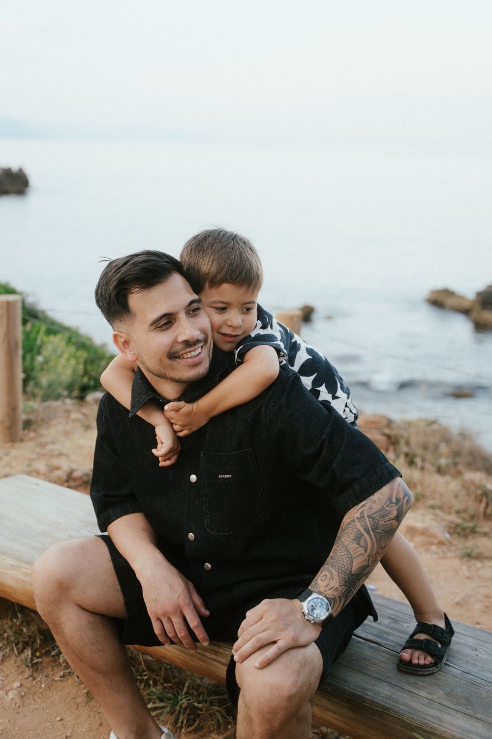 A smiling man with tattoos sits on a bench by the ocean while a young boy hugs him from behind.