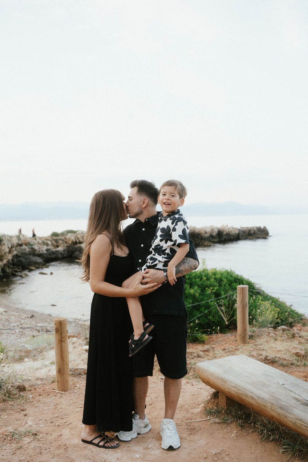 A couple shares a kiss while holding their smiling young son near a scenic coastal overlook.
