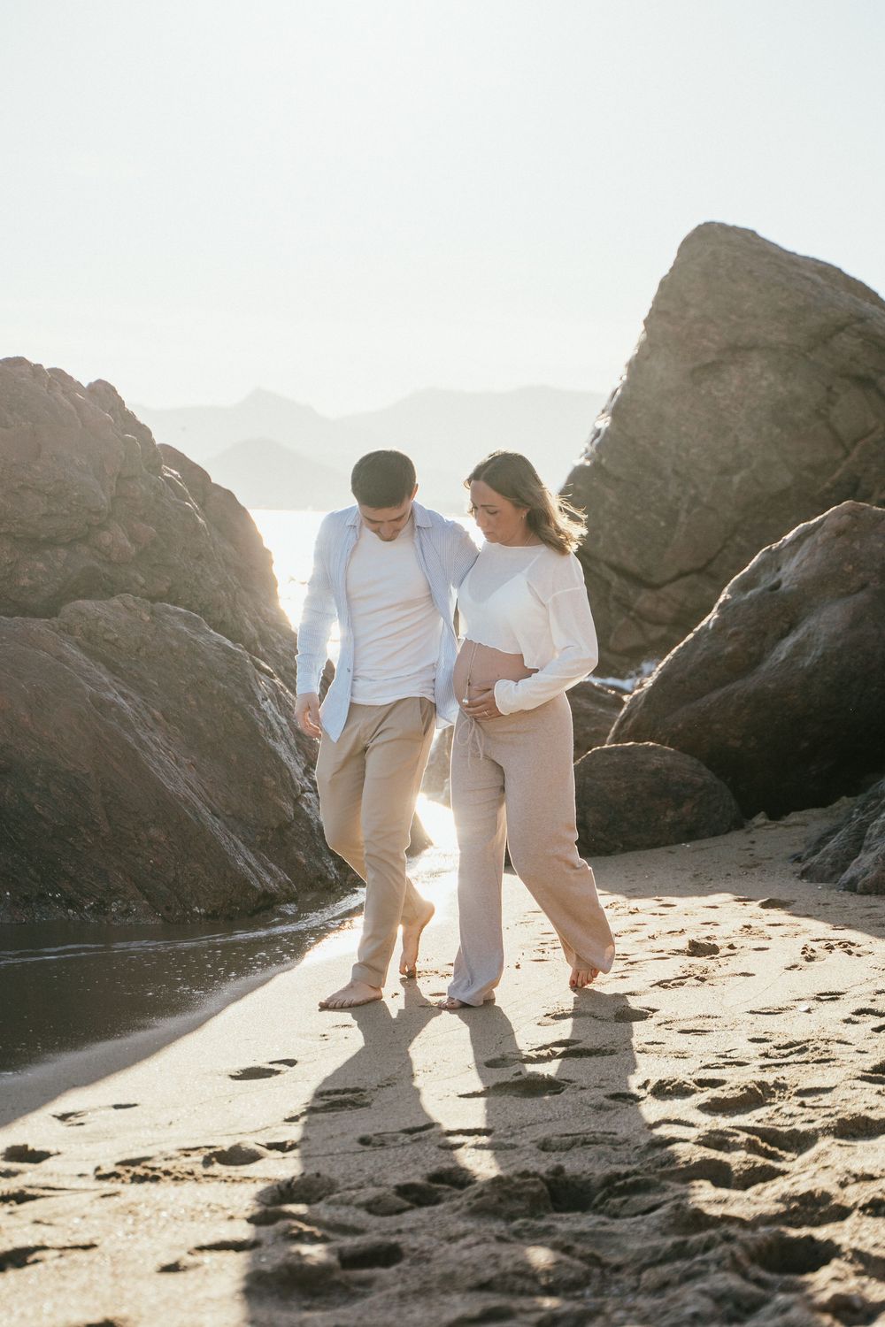 Couple walking barefoot on sandy beach between rocks, woman with baby bump, backlit by golden sunlight.