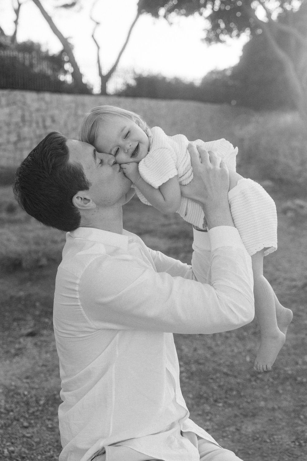 Black and white photo of a father lifting and kissing his laughing baby outdoors near trees.