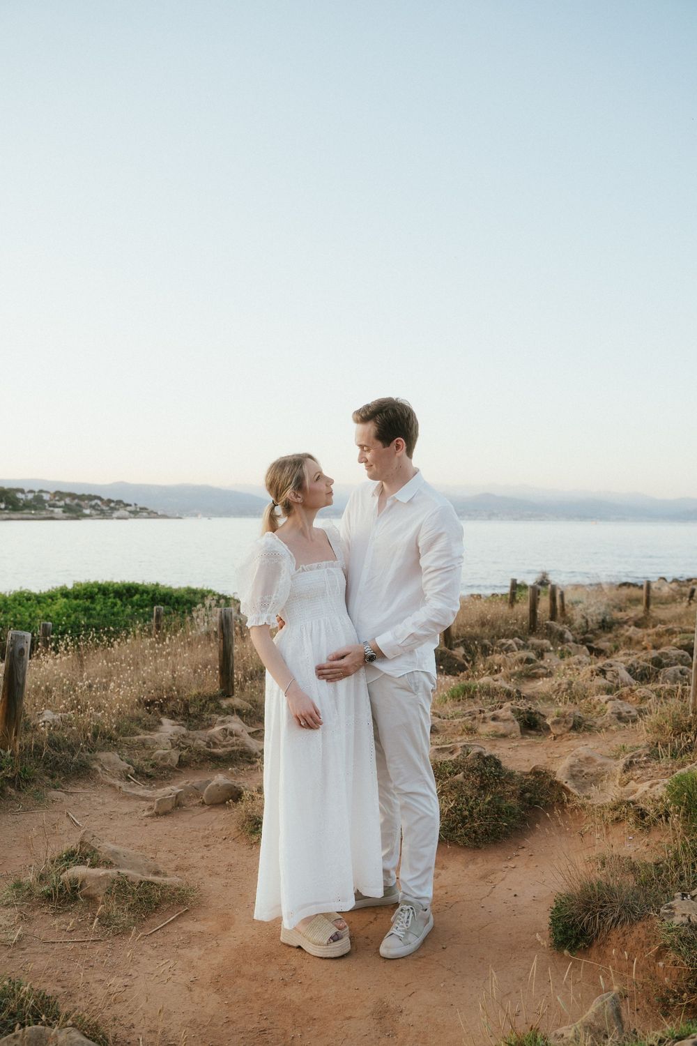 Couple in white outfits standing on a coastal path, woman in flowing dress with partner embracing her bump at sunset.