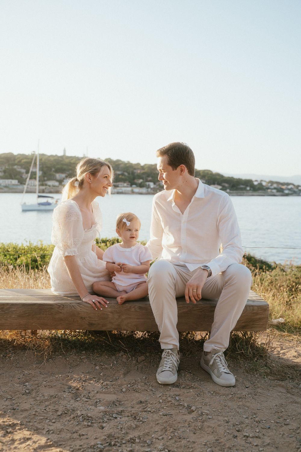A young family with a baby sits on a wooden bench by the seaside at golden hour, dressed in white and beige.