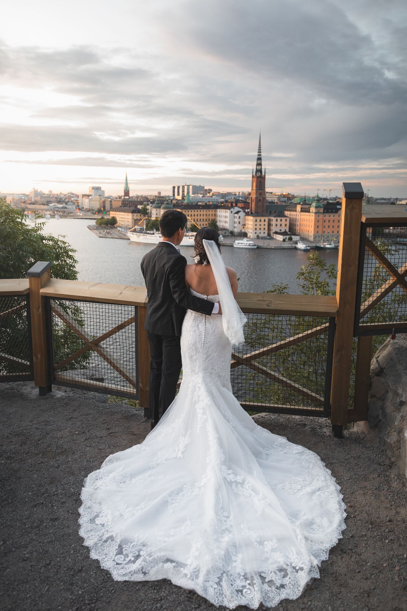 A couple in wedding attire gazes at the Stockholm cityscape from a scenic viewpoint at sunset.