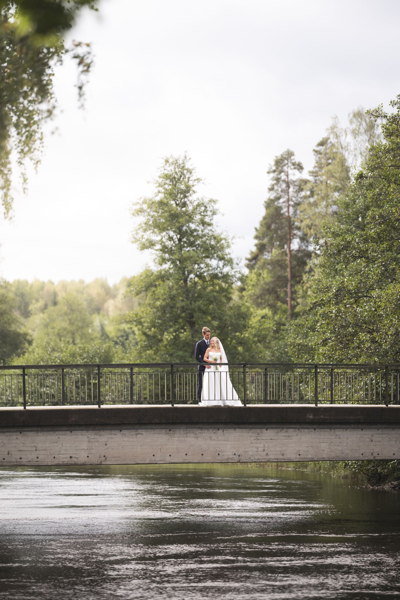 A couple shares a romantic moment on a wooden bridge over rippling water surrounded by lush green trees.