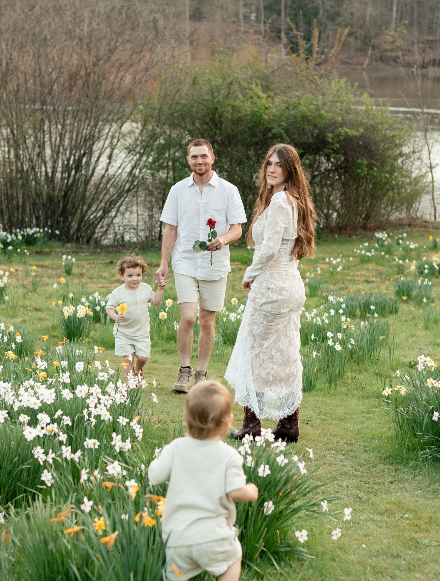 Two smiling young men with curly hair lean on a wooden railing outdoors, surrounded by lush green trees.
