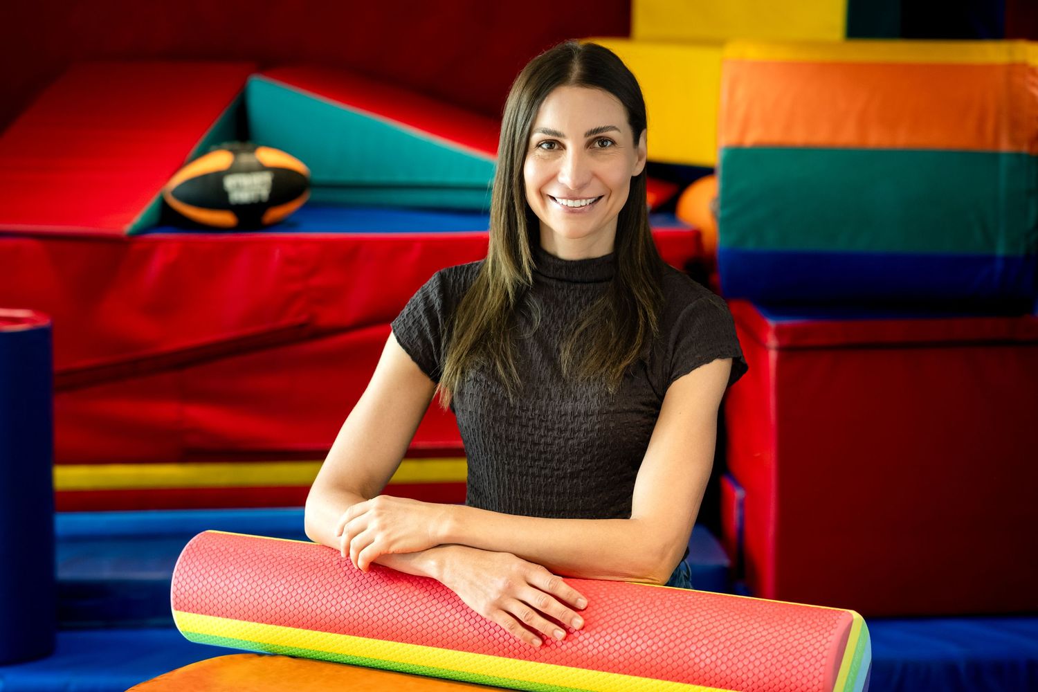 Smiling woman leaning on colorful foam roller in vibrant children's soft play area with red and blue padding.