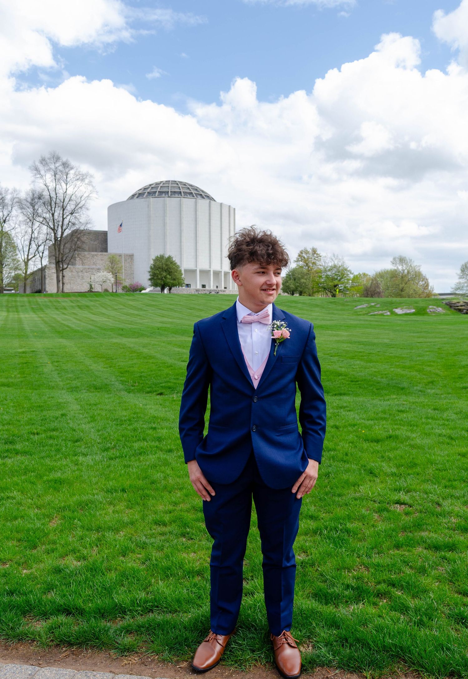 Young person in blue suit with floral boutonniere sitting on a rock outdoors among pine trees.