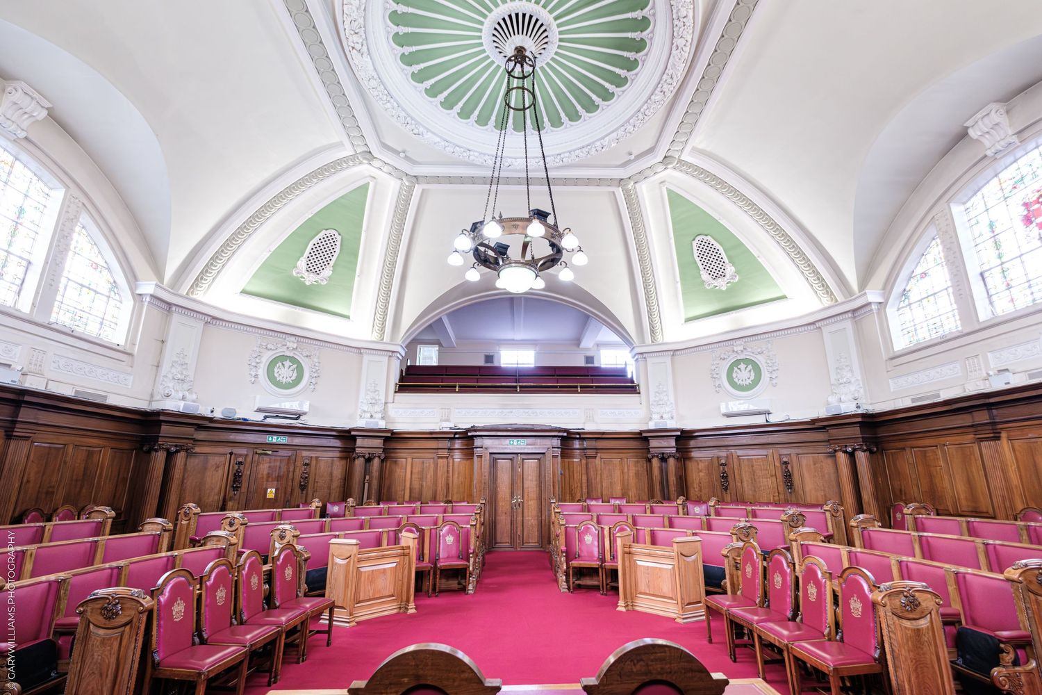 Elegant council chamber with pink upholstered chairs, ornate wooden paneling, and a decorative green domed ceiling with chandelier.