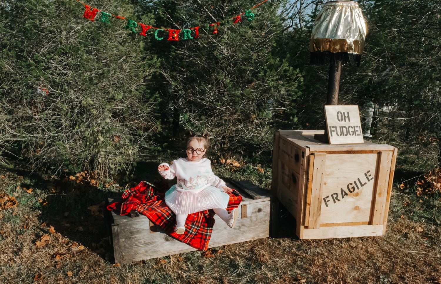 Baby in white dress with glasses sits in wooden crate beside FRAGILE box and lamp, Christmas tree background.