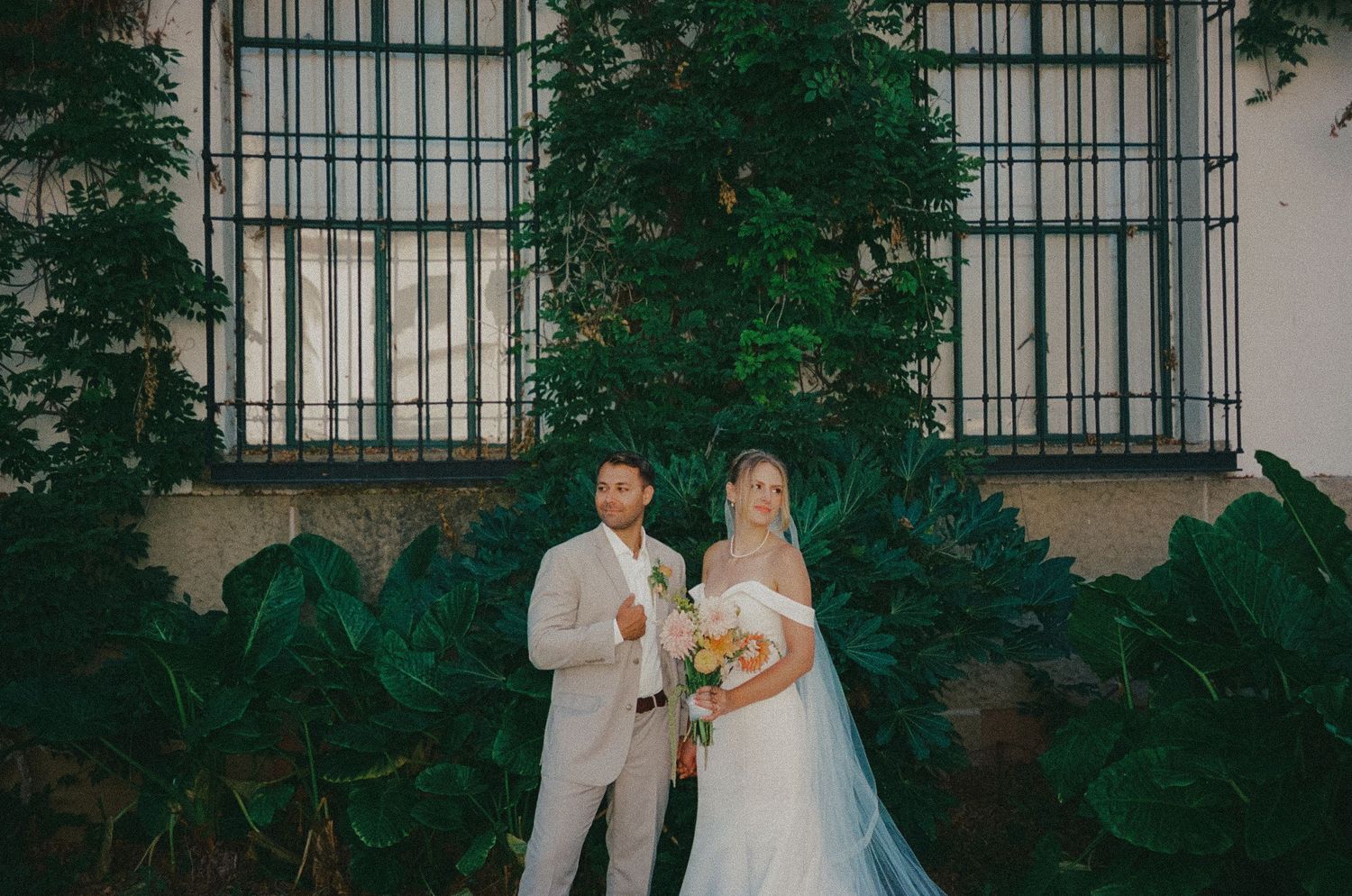 Bride and groom posing together in front of lush green foliage and iron window grill on their wedding day.
