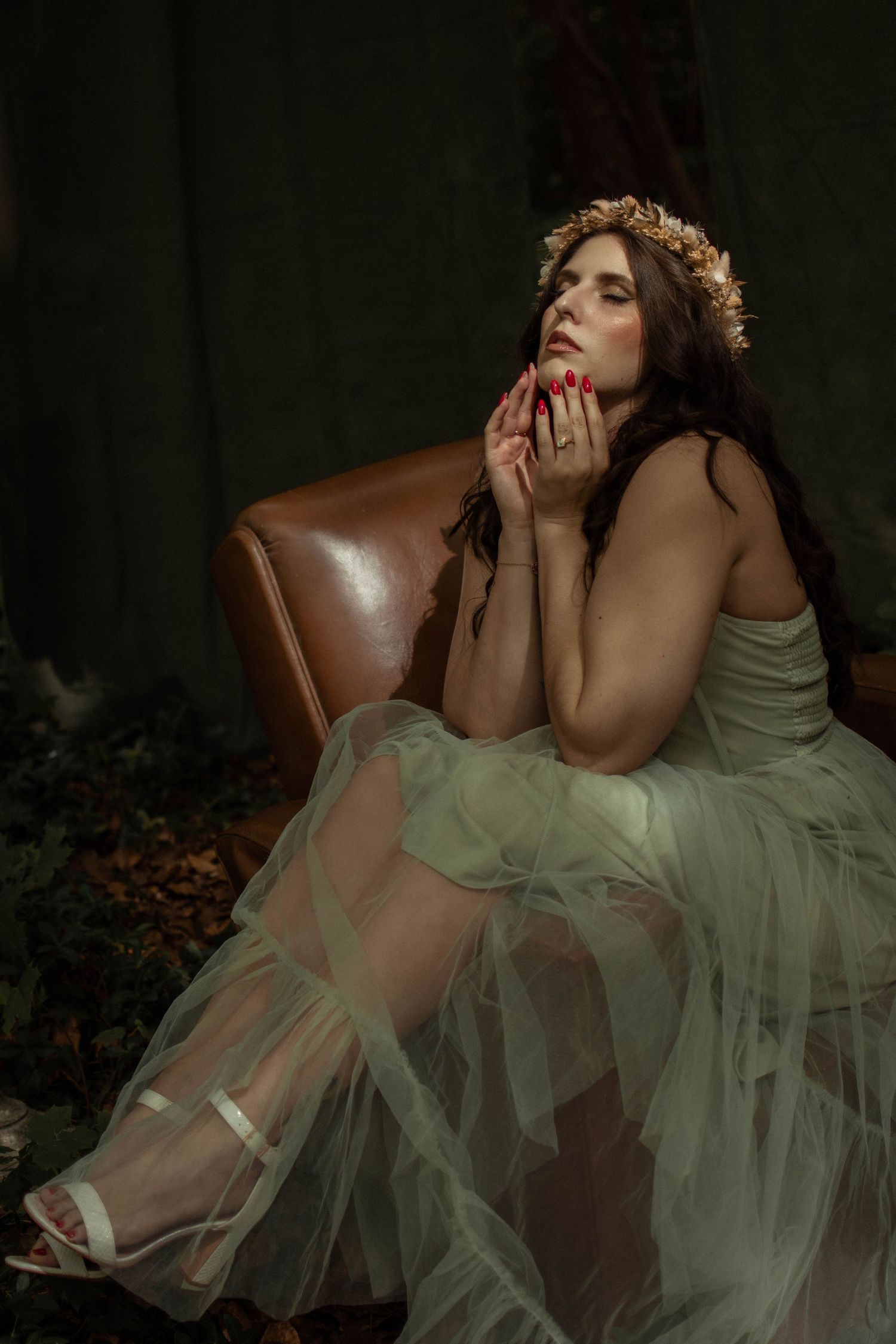 Woman in sage green tulle gown and floral crown sits gracefully in leather chair, gazing upward with red nails.