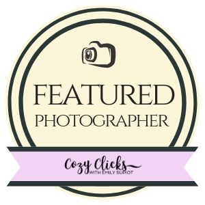 Cozy Clicks Featured Photographer badge with camera icon, cream background, and purple ribbon by Emily Slirot.