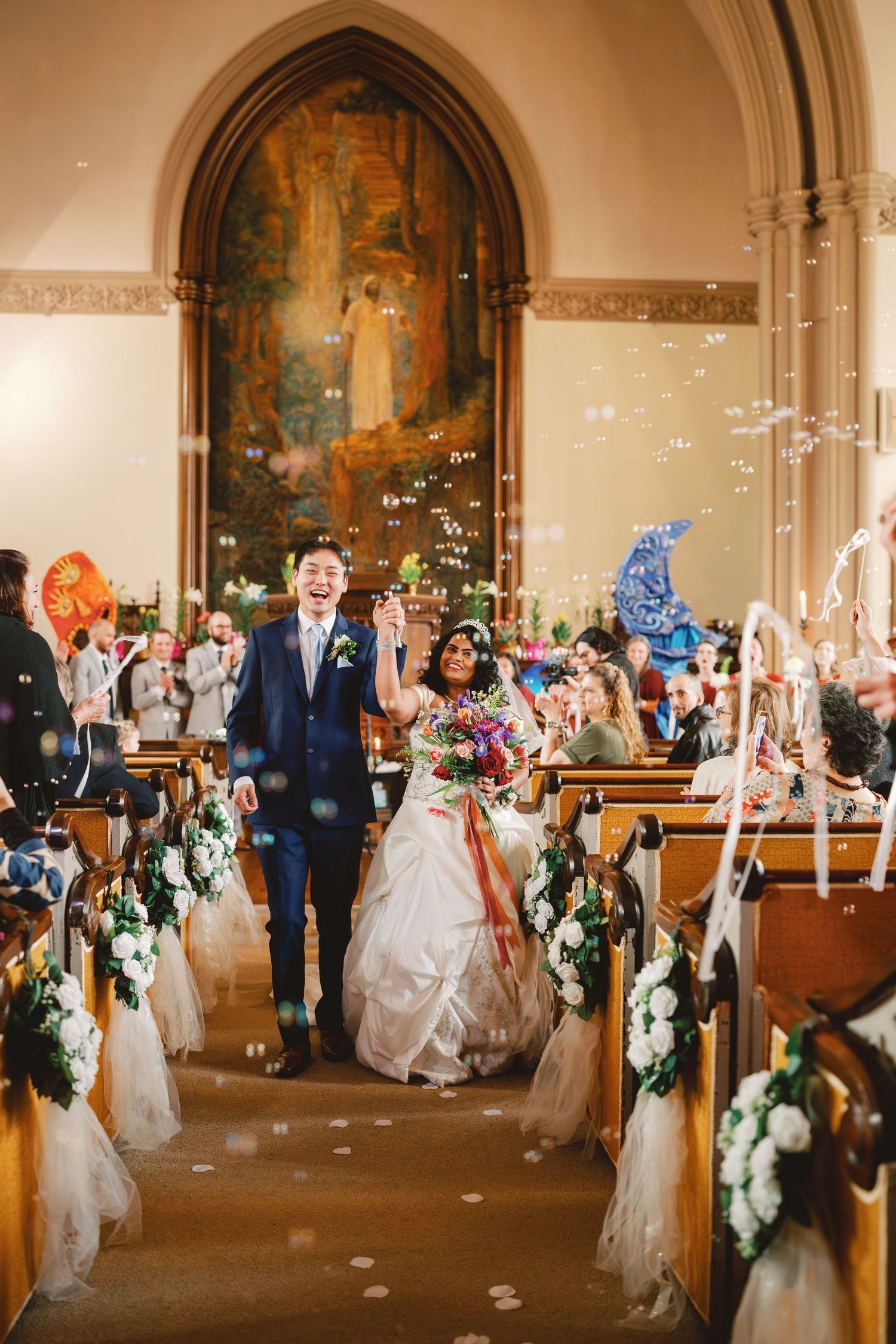 Joyful newlyweds walk down church aisle surrounded by bubbles, bride holding colorful bouquet, guests celebrating in wooden pew church.